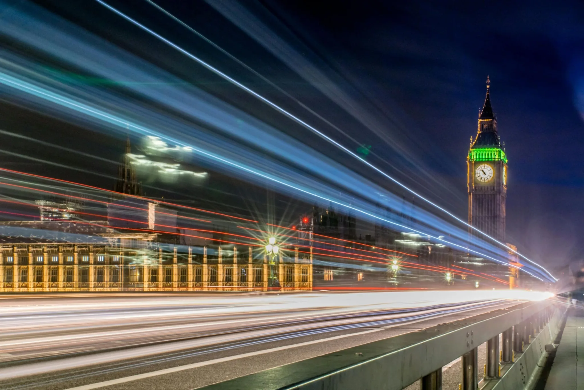 Night view of Big Ben and the Houses of Parliament in London, illuminated against a dark sky, with light trails from passing traffic showcasing energy and movement.